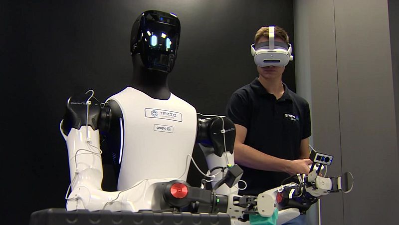Robot humanoide con logotipos "TEKIO" y "grupo i" junto a un hombre con visor VR y controlador, sugiriendo teleoperación en un entorno oscuro.