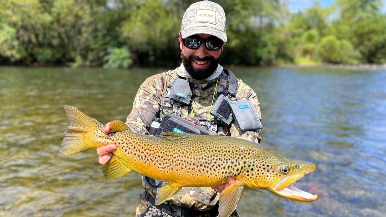 Campeón de pesca David Arcay sonríe con una gran trucha, luciendo equipo "ORCA" en un río rodeado de vegetación.