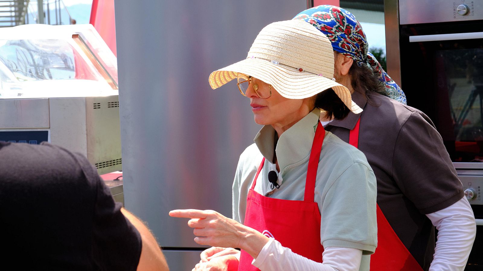 Paz Vega, con sombrero y delantal rojo, señala con el dedo en un evento de cocina. Otra persona con delantal está detrás.