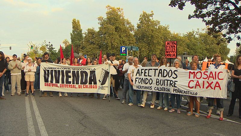 Manifestantes protestan en una calle urbana por la vivienda, exigiendo acceso gratuito y de calidad, y criticando a fondos buitre. Se ven pancartas con lemas como "ACABEMOS CON EL NEGOCIO DE LA VIVIENDA" y "AFECTADOS FONDO BUITRE".