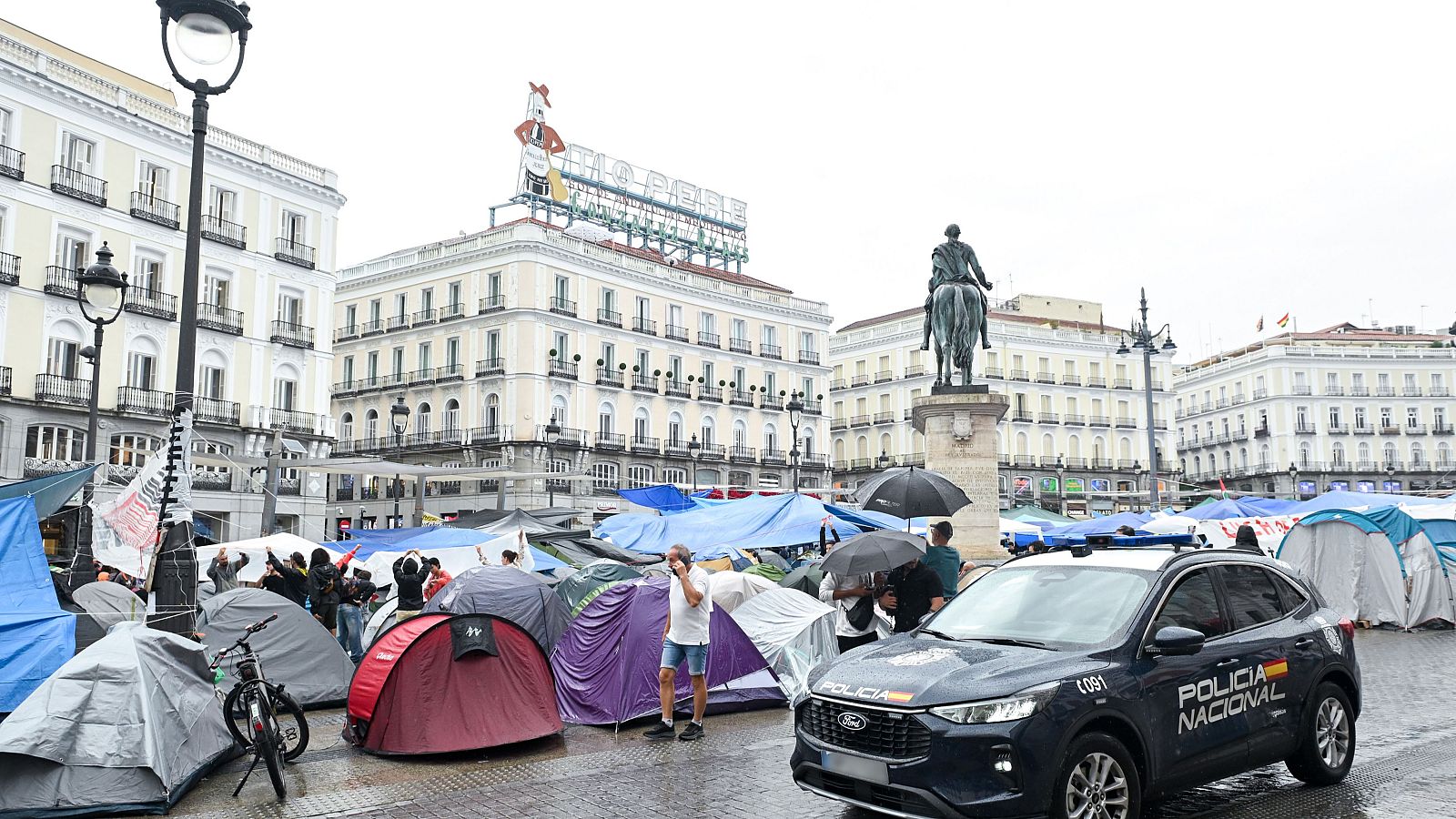 Vista de la Puerta del Sol con tiendas de campaña, estatua de Carlos III, edificio con cartel "TIO PEPE" y vehículo de Policía Nacional (091) bajo un día lluvioso.