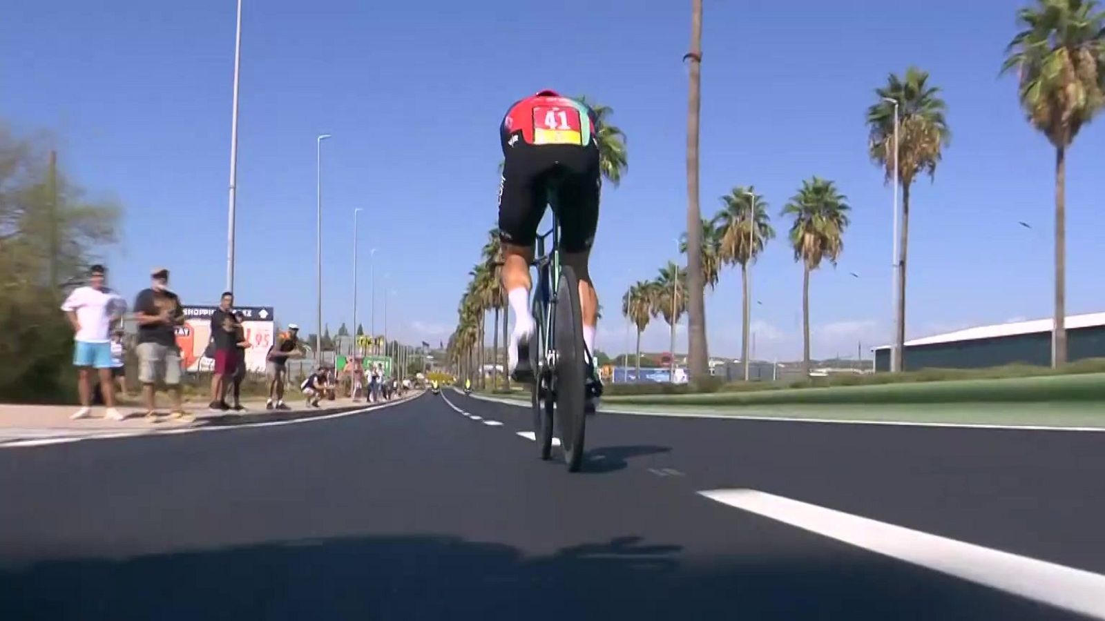 Ciclista con dorsal 41, maillot rojo y negro, avanza por carretera con palmeras. Espectadores observan. Cielo azul y soleado.