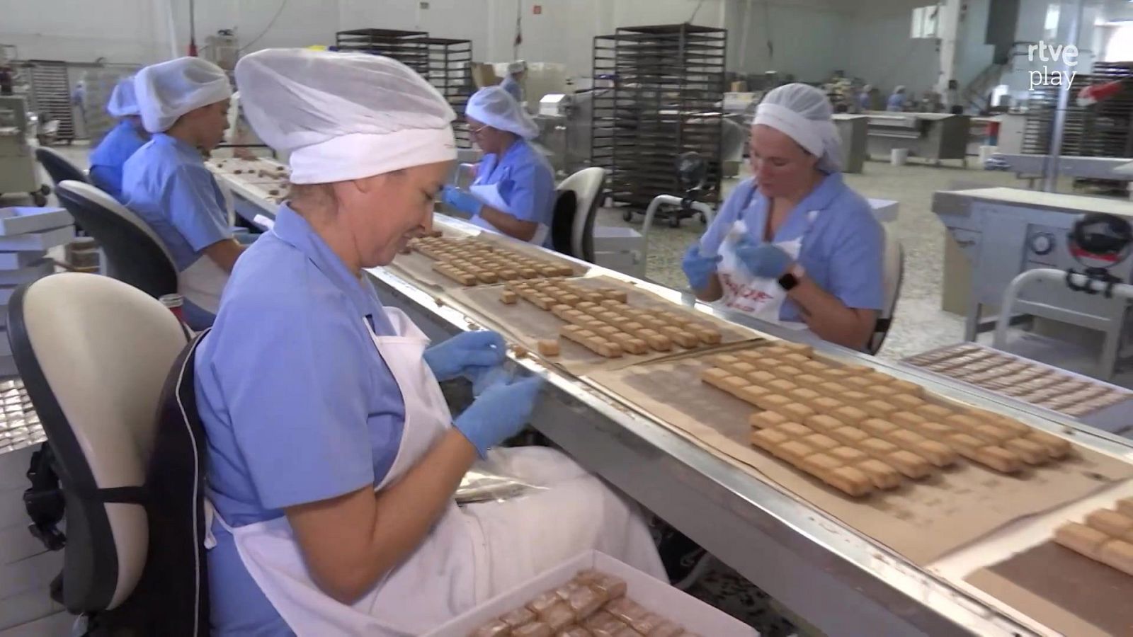 Trabajadoras con uniforme higiénico y guantes azules envasan polvorones en una cinta transportadora dentro de una fábrica de dulces navideños en Estepa, con el logo de RTVE Play en la esquina.