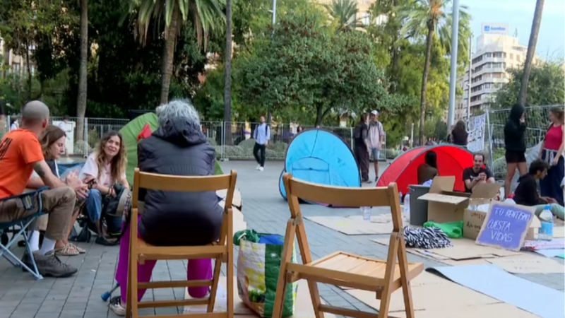 Acampada de protesta en una plaza con tiendas de campaña, personas conversando y un cartel morado en el suelo que dice "TU RENTABILIDAD CUESTA VIDAS".