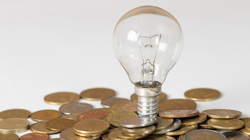 Una bombilla de vidrio transparente con filamento visible se apoya verticalmente sobre un montón de monedas de tonos dorados, plateados y cobrizos, sobre un fondo blanco.