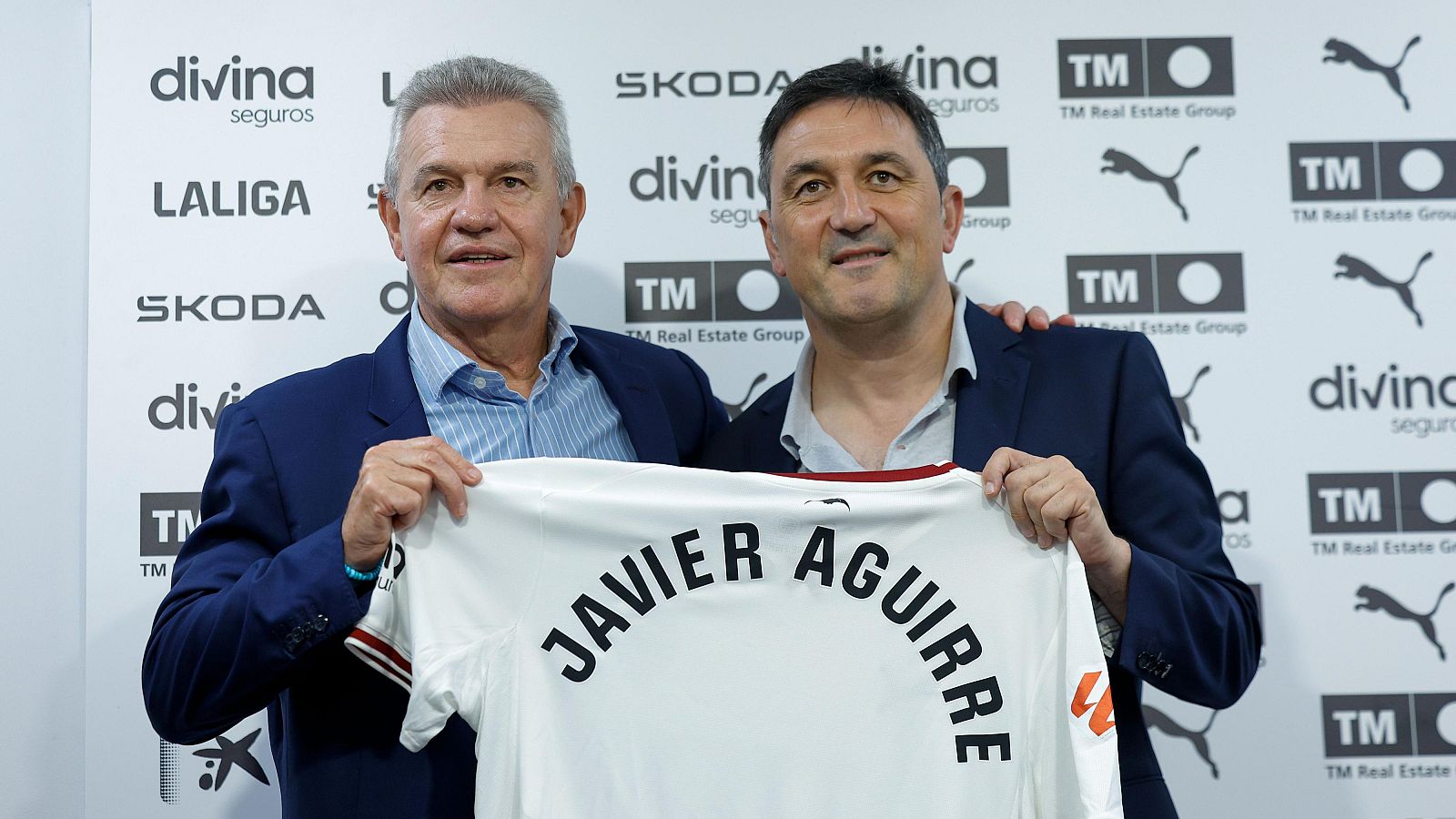 Javier Aguirre y Miguel Ángel Corona sonríen sosteniendo una camiseta blanca del Valencia CF con el nombre "JAVIER AGUIRRE" impreso, frente a un panel publicitario con logotipos.
