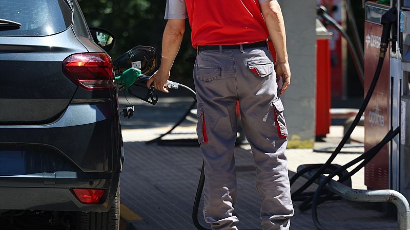 Un trabajador de espaldas con uniforme rojo y gris reposta gasolina en un coche oscuro usando una manguera verde en una estación de servicio Cepsa.