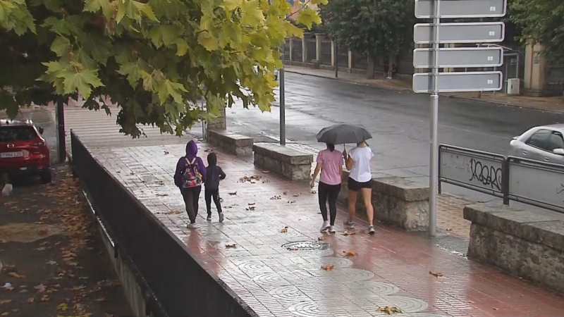 Peatones caminan bajo la lluvia por una acera mojada y con hojas otoñales, algunos protegidos por un paraguas negro, en una calle de Extremadura durante un día de fuertes precipitaciones.