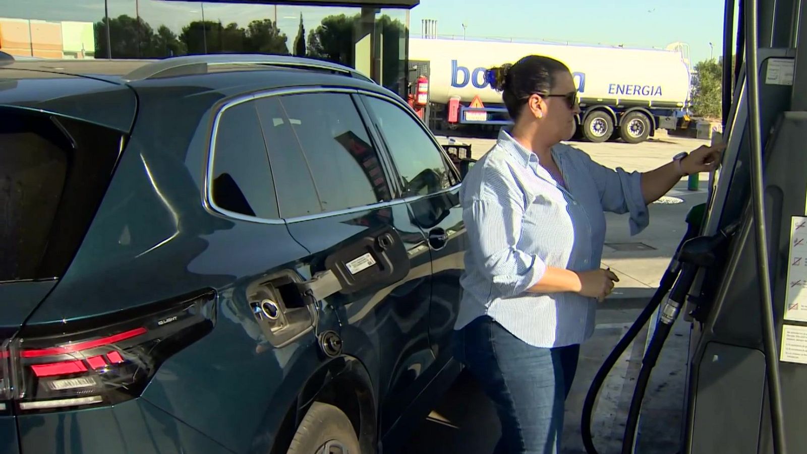 Una mujer con gafas de sol paga en el surtidor de una gasolinera junto a su SUV azul, con un camión cisterna de bonÀrea ENERGIA al fondo.