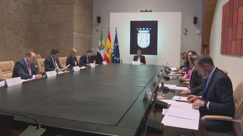 María Guardiola preside la reunión del Consejo de Gobierno de la Junta de Extremadura junto a sus consejeros en una sala con banderas oficiales y una pared de piedra rústica.