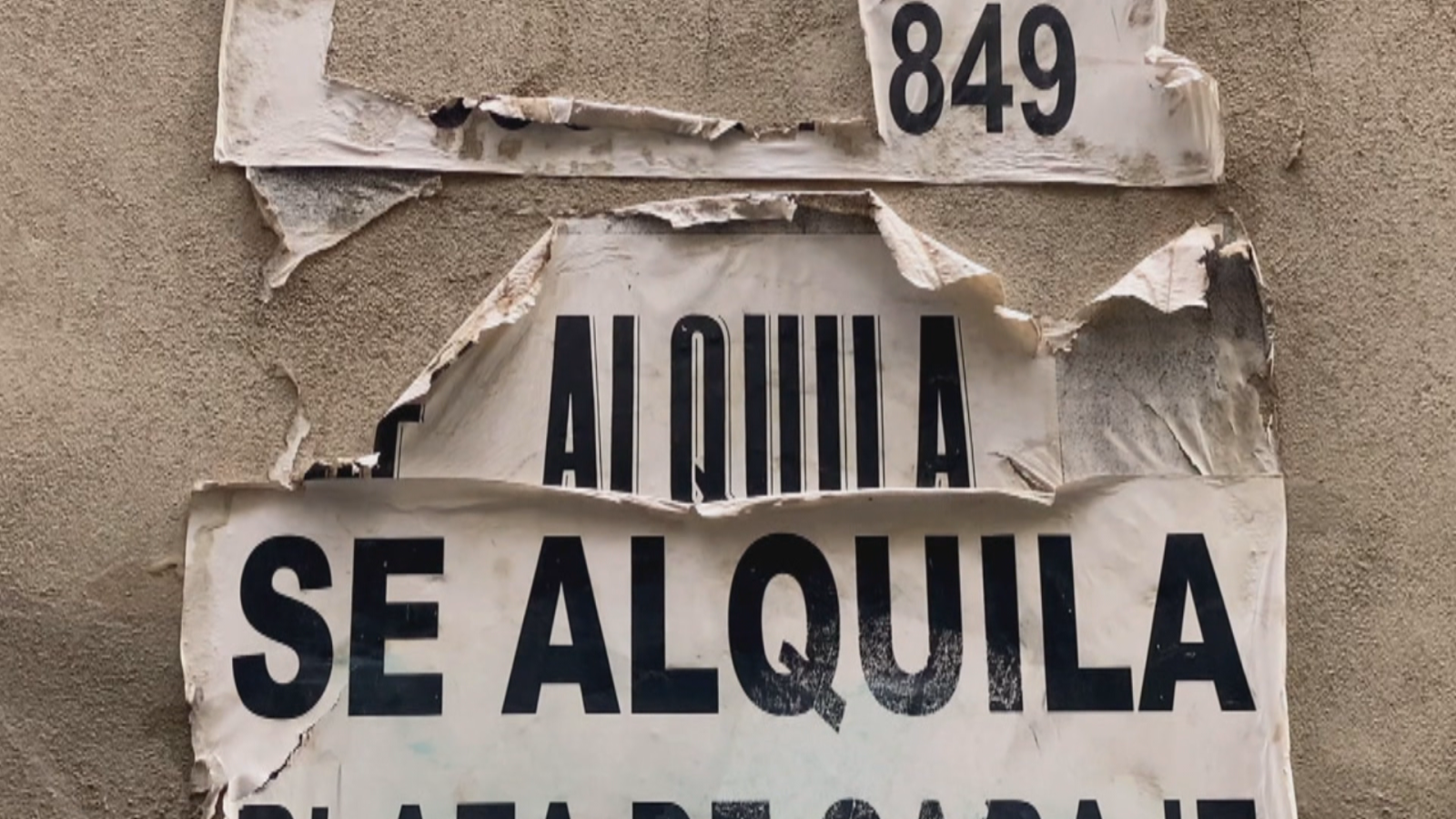Varios carteles superpuestos y rotos sobre una pared gris muestran los textos "849", "ALQUILA" y "SE ALQUILA PLAZA DE GARAJE" en letras negras.