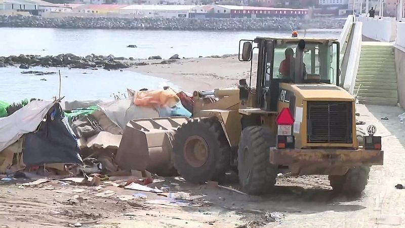 Pala cargadora amarilla CAT retira escombros en una playa de Ceuta, frente a naves industriales al fondo, destacando el cartel "ALMACENES SAN PABLO".