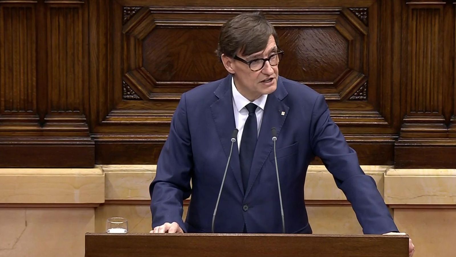 Salvador Illa, con traje azul marino y gafas, interviene con semblante serio desde el atril de madera del Parlament de Catalunya.