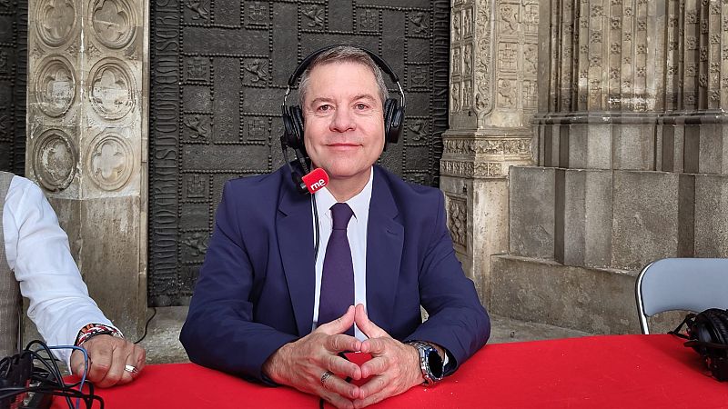 Emiliano García-Page, con traje azul y corbata, sonríe usando auriculares de RNE durante una entrevista al aire libre frente a una gran puerta monumental de piedra.