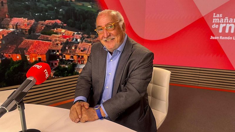 Juan Ramón Lucas, con gafas amarillas y traje gris, sonríe en el estudio de "Las mañanas de RNE" frente a un micrófono rojo y un panel con su nombre.