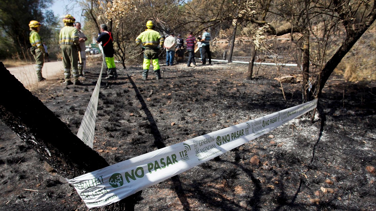 Cinta de la Generalitat Valenciana con el texto "NO PASAR" en primer plano de un bosque quemado, con bomberos y civiles evaluando los daños al fondo.