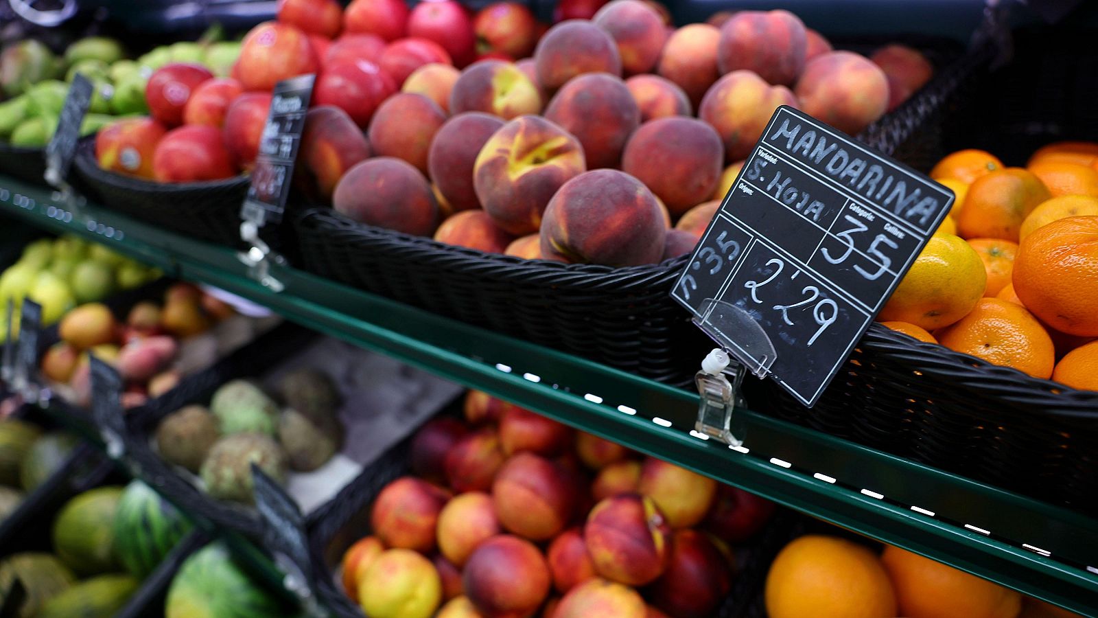 Primer plano de un cartel de pizarra en una frutería que indica "MANDARINA", "S. Hoja", "Calibre 35" y el precio de "2'29", rodeado de mandarinas y otras frutas frescas.