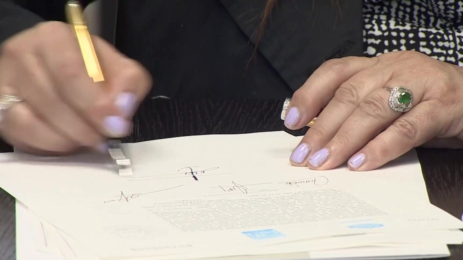 Primer plano de las manos de una mujer con un anillo de piedra verde firmando un documento oficial con un bolígrafo dorado sobre una mesa de madera.