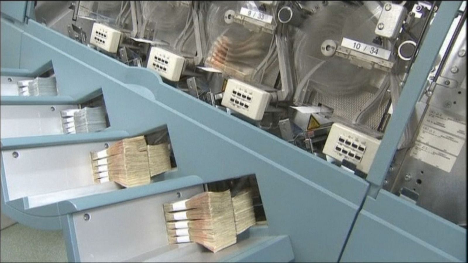Detalle de una máquina industrial clasificadora con fajos de billetes de 50 euros y paneles de control con las etiquetas "9 / 33" y "10 / 34".