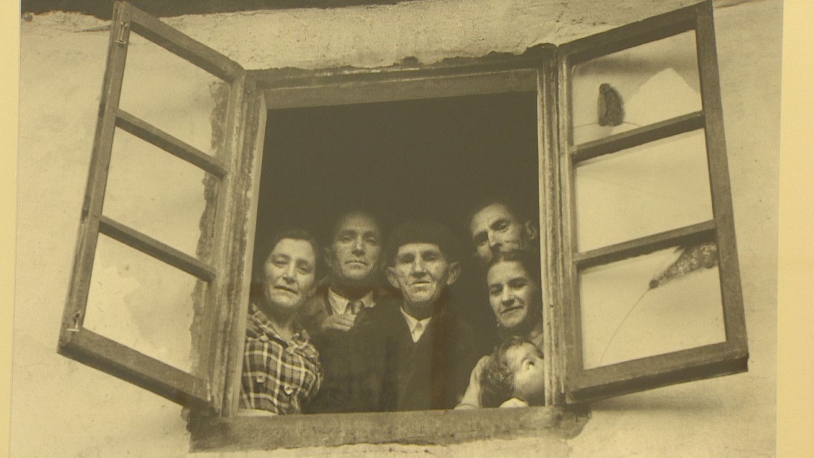 Retrato en sepia de una familia rural de mediados del siglo XX, con seis personas de varias generaciones asomadas a una ventana de madera abierta en una pared de adobe.
