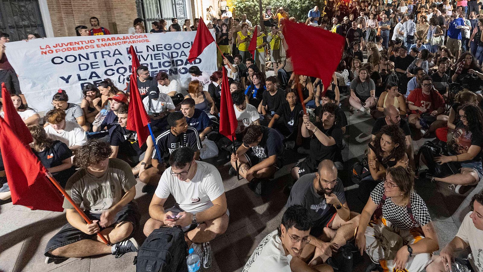 Jóvenes sentados en una protesta nocturna con banderas rojas y una pancarta que reza: 'LA JUVENTUD TRABAJADORA NO PODEMOS EMANCIPARNOS'.