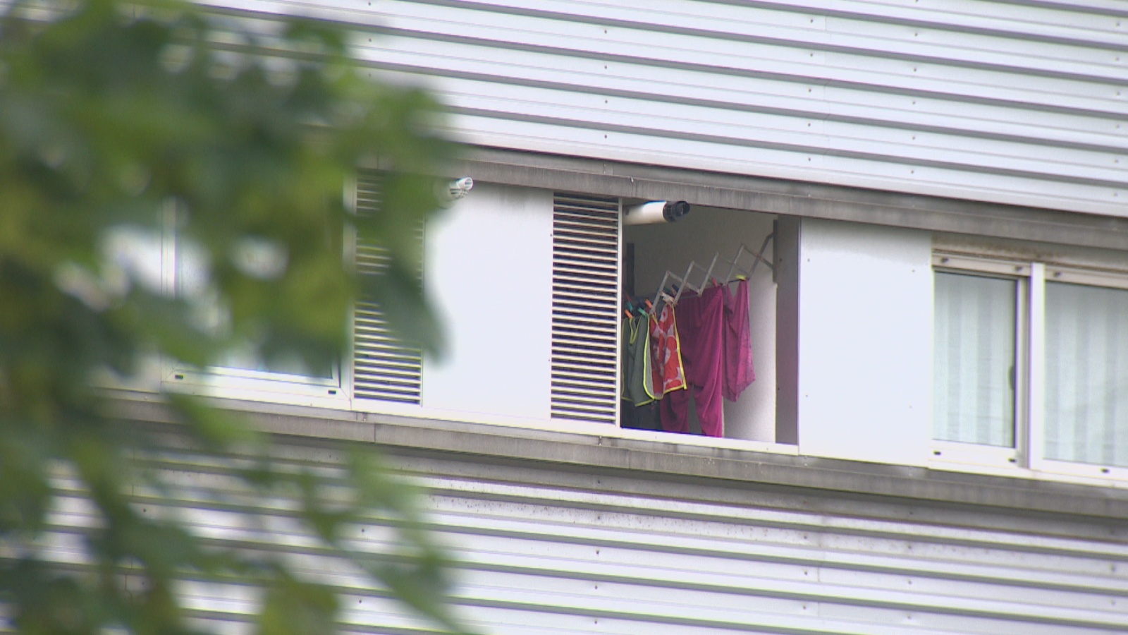 Fachada de un edificio moderno de paneles grises con un balcón empotrado donde hay ropa tendida de colores y ventanas con marcos blancos a la derecha.