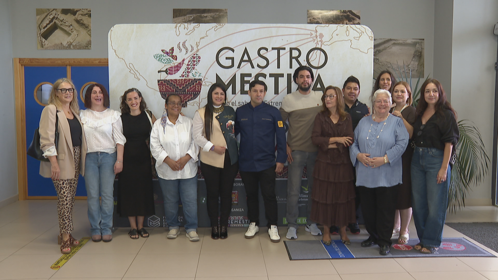 Trece personas posan sonrientes en un vestíbulo frente a un cartel de "GASTRO MESTIZA ...el sabor de Extremadura" decorado con un mortero e ingredientes culinarios.