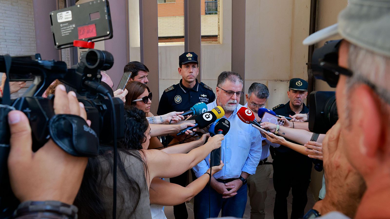 Un hombre con gafas declara ante periodistas con micrófonos de À Punt, Cadena SER y COPE, con agentes de la Policía Local de fondo frente a un edificio.