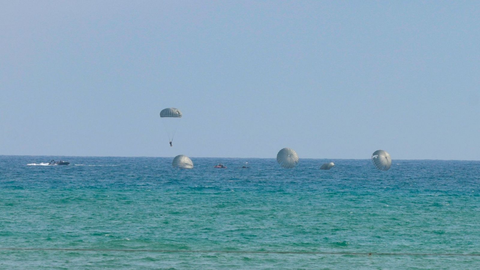 Un paracaidista militar desciende sobre el mar turquesa junto a lanchas de apoyo y otros paracaídas flotando en el agua durante unas maniobras del Ejército del Aire.