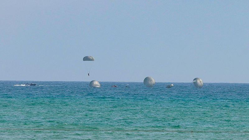 Un paracaidista militar desciende sobre el mar turquesa junto a lanchas de apoyo y otros paracaídas flotando en el agua durante unas maniobras del Ejército del Aire.