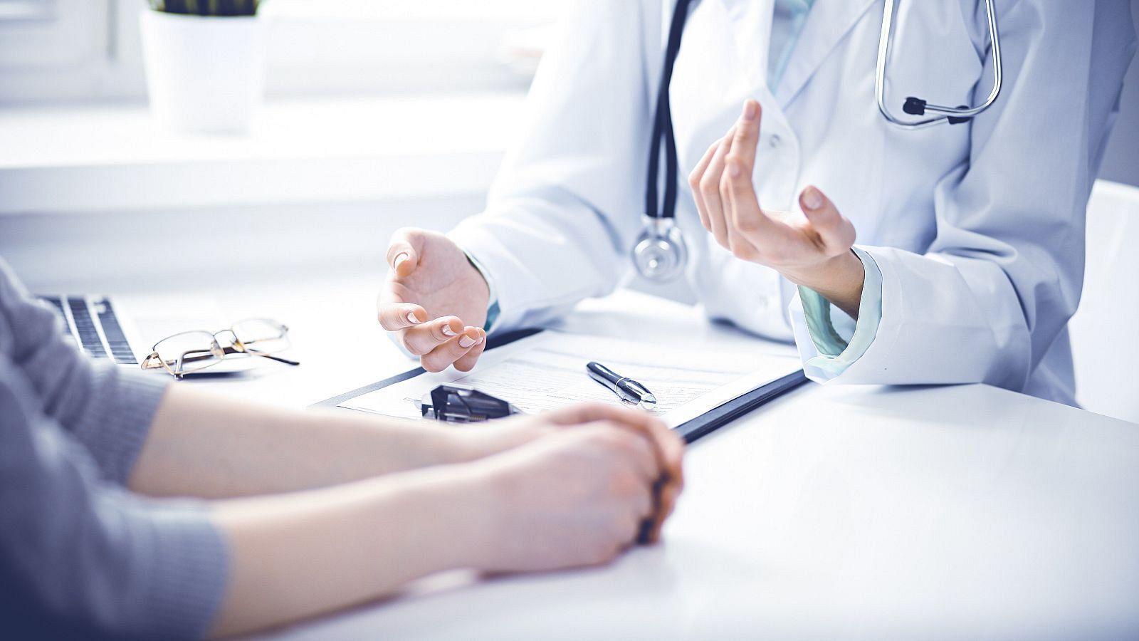 Un médico, con bata y estetoscopio, conversa con un paciente en un consultorio. En la mesa hay un portapapeles, un bolígrafo, un ordenador portátil y unas gafas, sugiriendo una consulta médica.