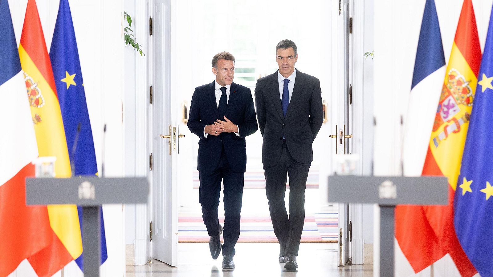 Pedro Sánchez y Emmanuel Macron caminan sonrientes en un acto oficial entre las banderas de España, Francia y la Unión Europea, con atriles del Gobierno de España en primer plano.