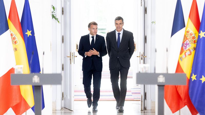Pedro Sánchez y Emmanuel Macron caminan sonrientes en un acto oficial entre las banderas de España, Francia y la Unión Europea, con atriles del Gobierno de España en primer plano.
