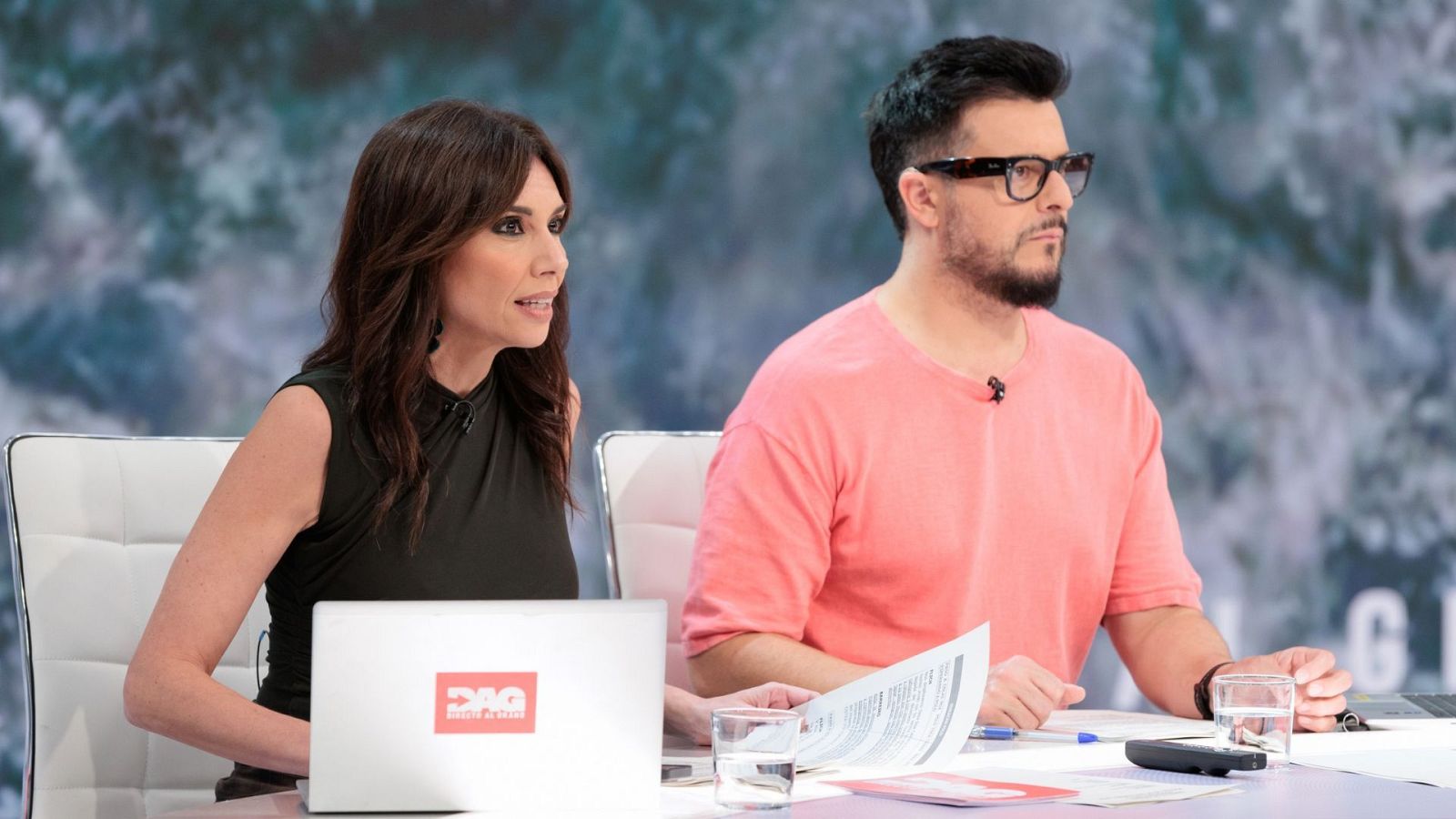 Los presentadores Raquel Tejero y Luis Losada Pescador en un plató de televisión tras una mesa con un portátil con el logo 'DAG Directo al grano', papeles y vasos de agua.