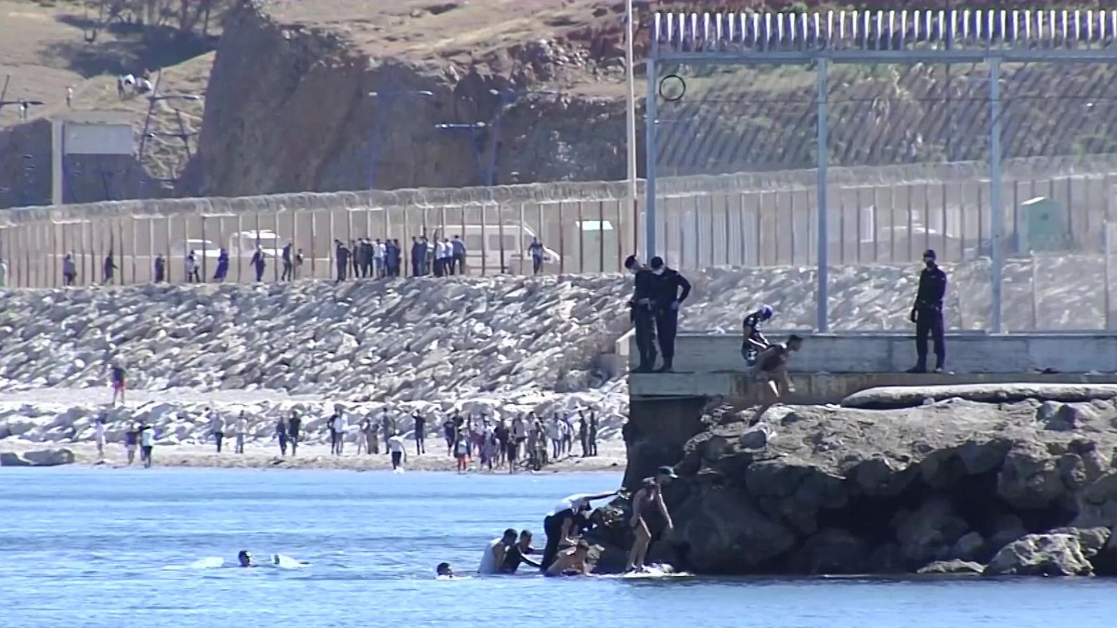 Personas nadando e intentando subir a un espigón de rocas en Ceuta, vigiladas por agentes de seguridad junto a la valla fronteriza.