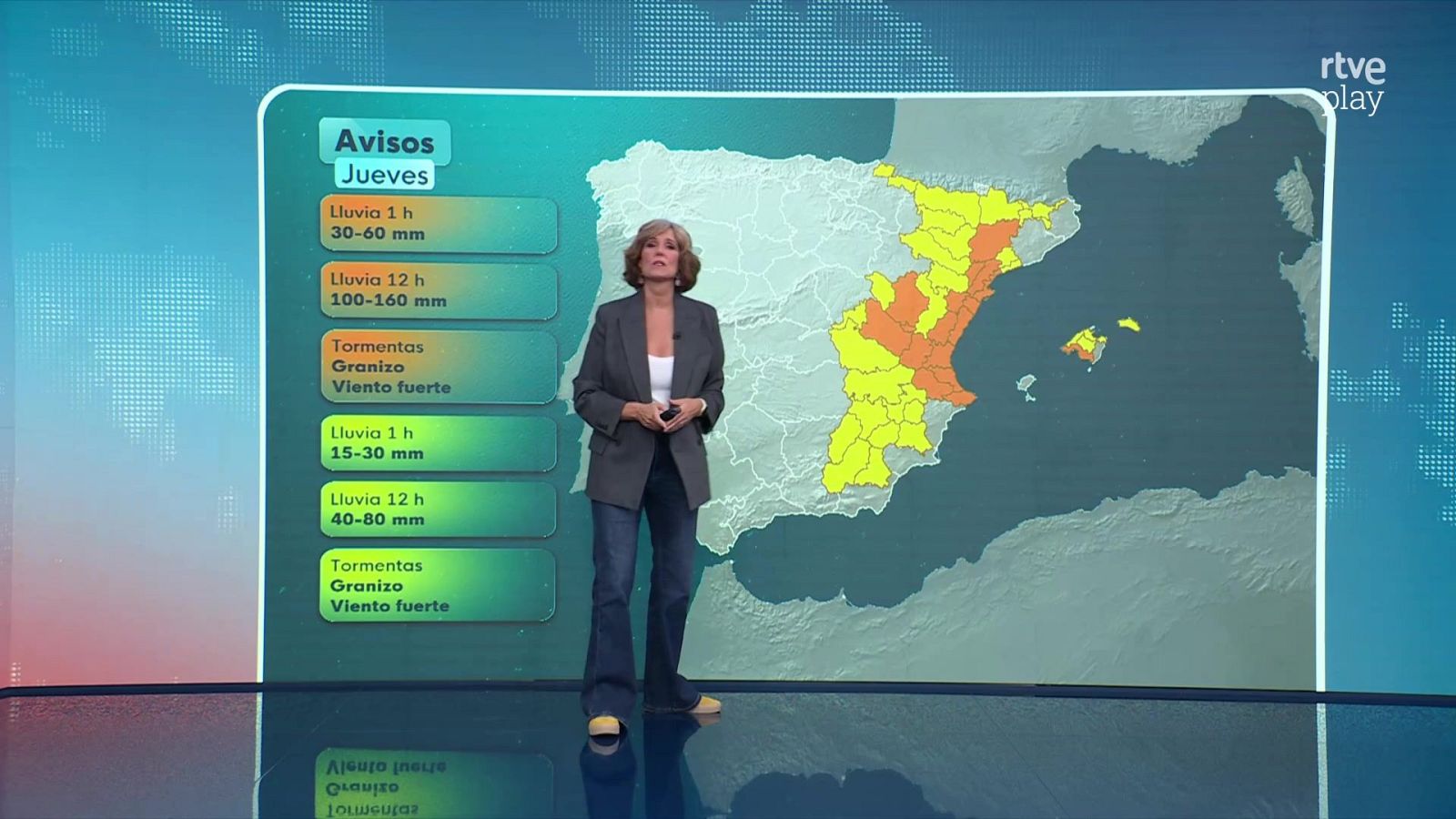 La presentadora Ana de Roque, vestida con chaqueta gris y vaqueros, expone en el plató de RTVE un mapa de España con avisos meteorológicos por lluvias y tormentas para el jueves.
