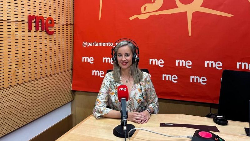 La periodista Diana Arias sonríe en un estudio de RNE, con auriculares y frente a un micrófono con el logo de la emisora sobre un fondo rojo decorado con @parlamento_rne.