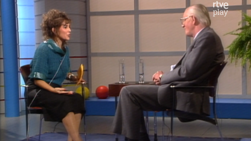 Julia Otero entrevista a Tom Sharpe en un plató de RTVE de los años 80, sentados frente a frente junto a una mesa con vasos de agua.