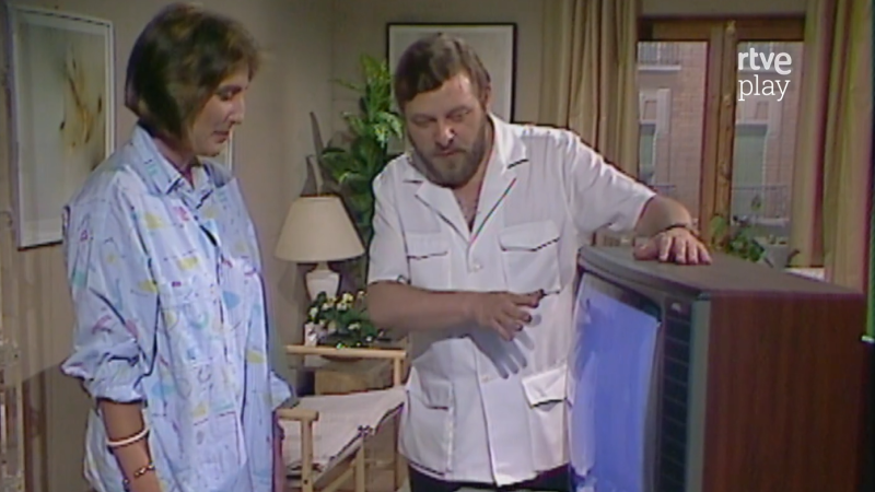 Un hombre con barba señala la pantalla azul de un televisor antiguo de madera junto a una mujer que lo observa, en un plató decorado como un salón con el logo de rtve play.