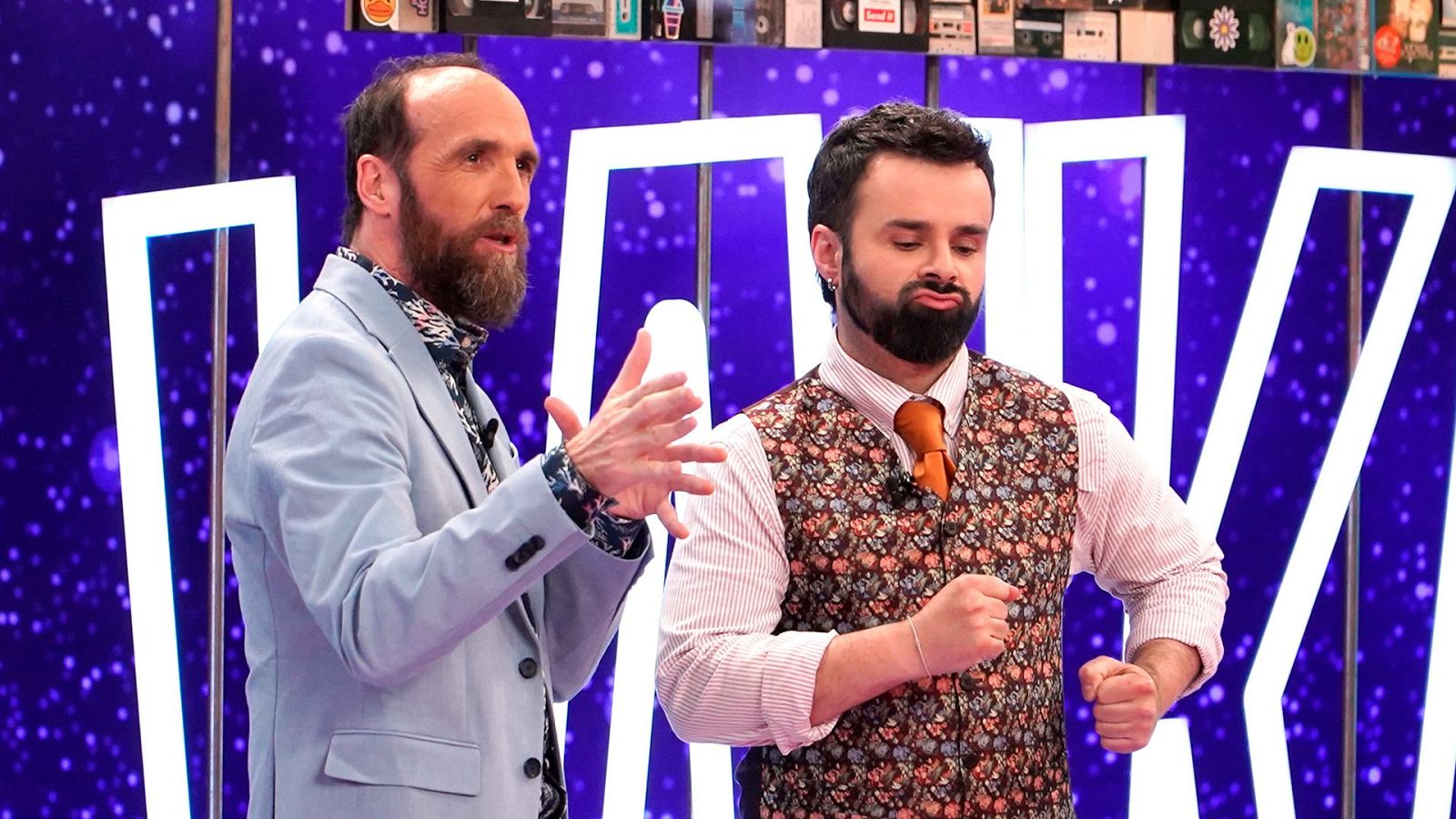 Eduardo Sáenz de Cabezón y Ricardo Moure gesticulan divertidos en el plató de "Órbita Laika", decorado con letras iluminadas y elementos retro de los noventa sobre un fondo violeta.