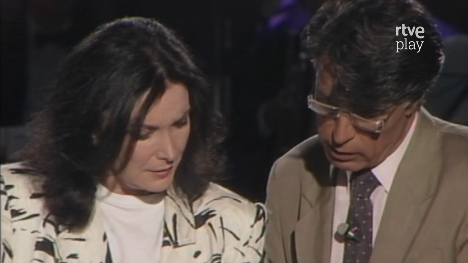 Nuria Espert, con chaqueta estampada, y Jesús Hermida, con traje y gafas, miran hacia abajo en un plató de televisión de RTVE Play.