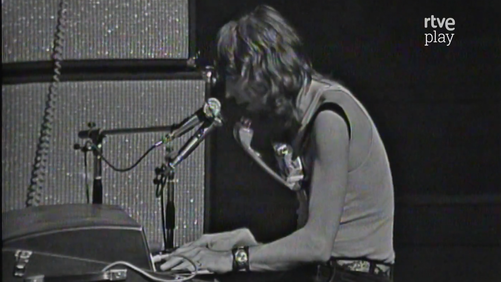 John Mayall, de perfil y con camiseta sin mangas, toca un teclado y lleva una armónica al cuello en una actuación en blanco y negro con el logo de "rtve play".
