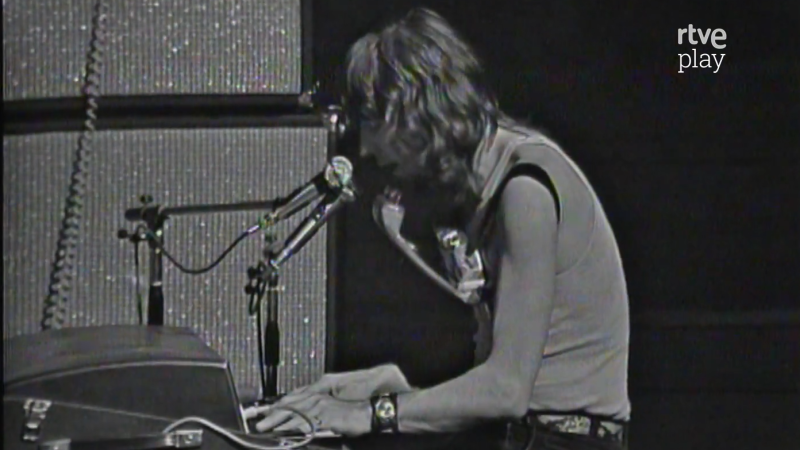 John Mayall, de perfil y con camiseta sin mangas, toca un teclado y lleva una armónica al cuello en una actuación en blanco y negro con el logo de "rtve play".