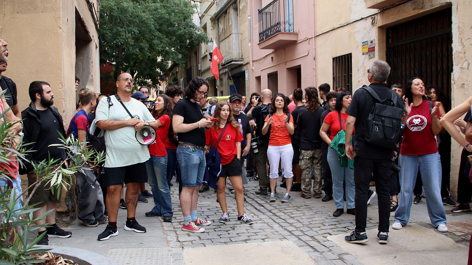 Vecinos reunidos en una calle peatonal adoquinada participan en una protesta, con un hombre sosteniendo un megáfono y otros aplaudiendo frente a edificios antiguos.