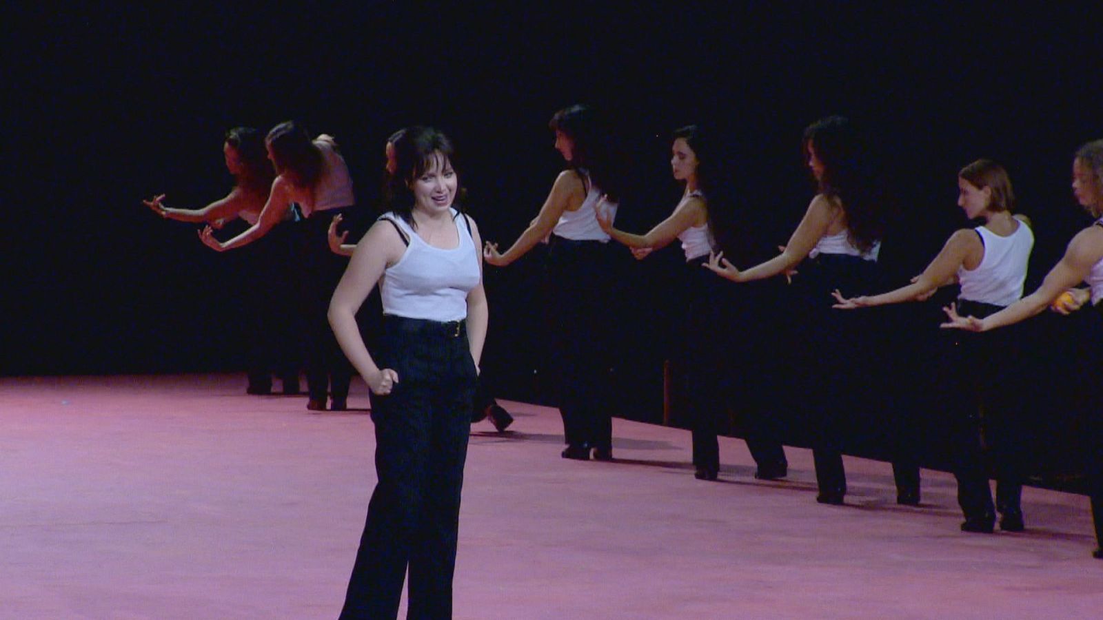 Artistas con camisetas blancas y pantalones negros realizan una coreografía estilizada sobre un escenario de suelo rosa y fondo oscuro en una representación de la ópera Carmen.