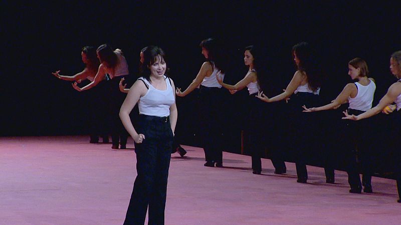 Artistas con camisetas blancas y pantalones negros realizan una coreografía estilizada sobre un escenario de suelo rosa y fondo oscuro en una representación de la ópera Carmen.
