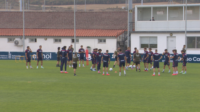 Jugadores del Osasuna entrenan en Tajonar con ropa azul y fucsia, rodeados de balones y vallas publicitarias con logos de "Gobierno de Navarra", "Enhol" y "Hyundai".