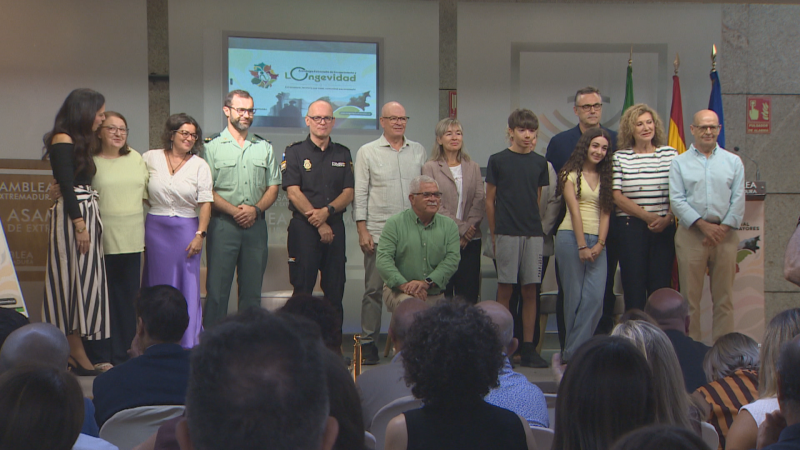 Grupo de personas, incluyendo agentes de la Guardia Civil y Policía Nacional, posa en el escenario de la Asamblea de Extremadura bajo una pantalla con la palabra "Longevidad".