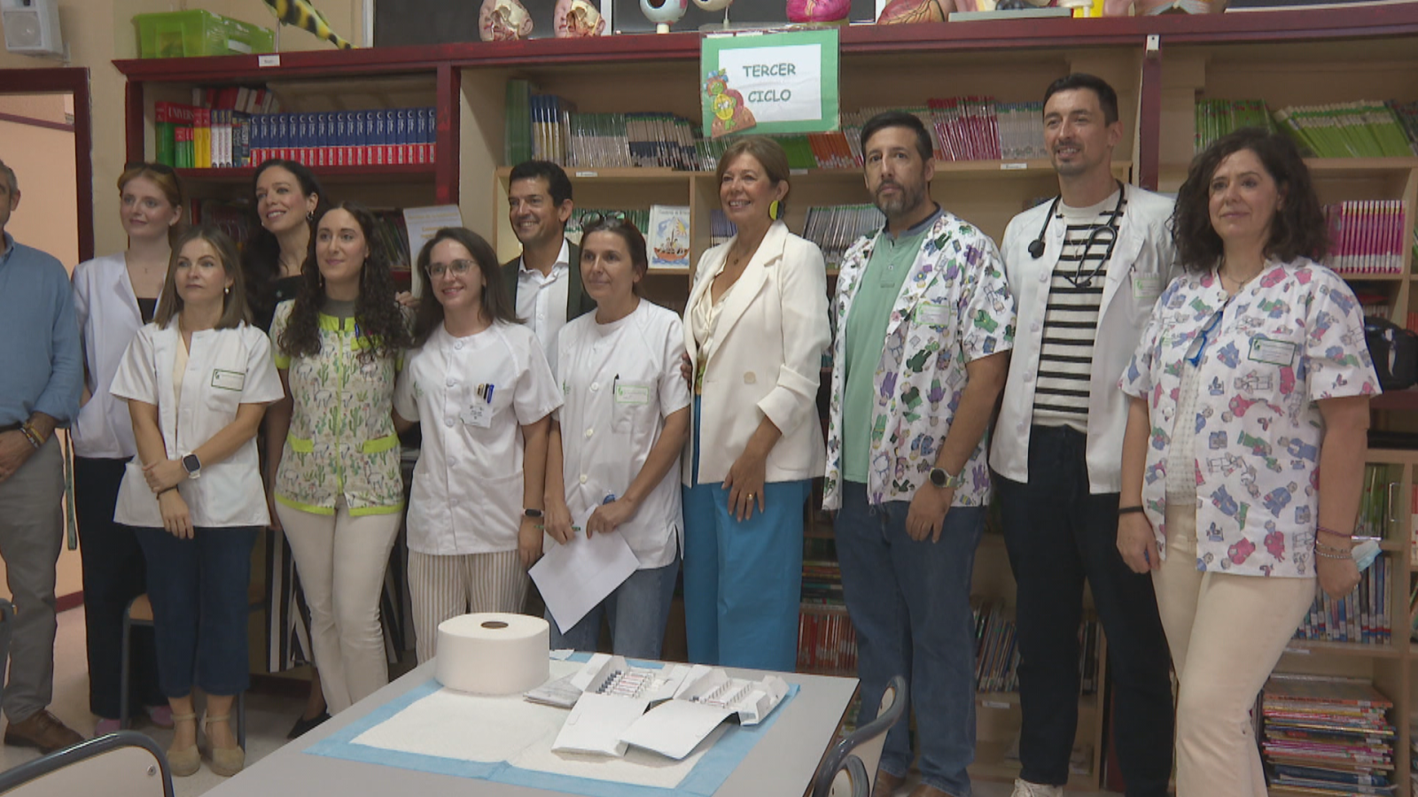 María Guardiola y Sara García Espada posan junto a personal sanitario en una biblioteca escolar con un cartel de "TERCER CICLO" y material de vacunación en primer plano.
