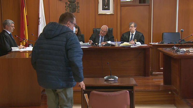 Un acusado de espaldas comparece ante un tribunal de tres magistrados en una sala de vistas de Cantabria, con las banderas oficiales y el escudo constitucional de fondo.